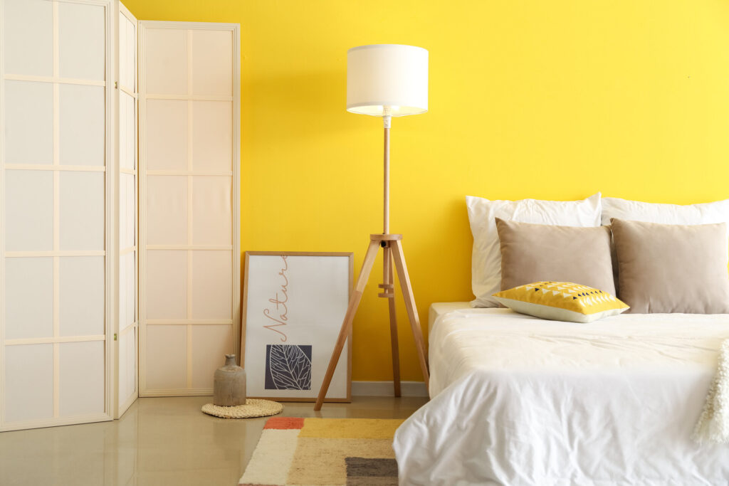 Stilvolles Schlafzimmer mit bequemem Bett, Stehlampe und Paravent vor einer gelben Wand 