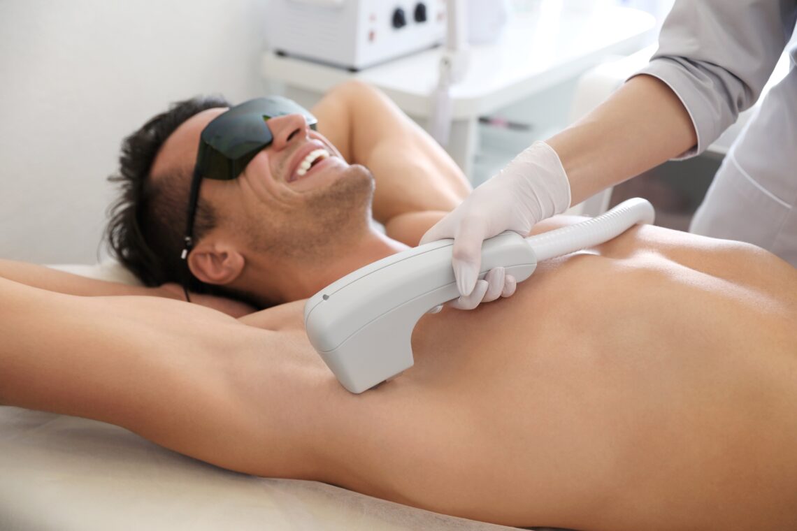Ein Mann unterzieht sich einer Laser-Haarentfernung unter den Achseln in einer Klinik, während er eine Schutzbrille trägt und lächelt