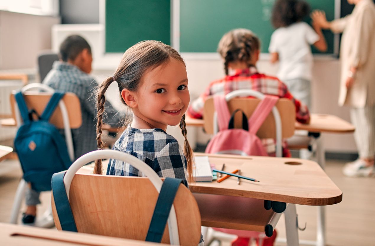 Laechelndes Maedchen mit Zopfen in der grundschule berlin, sitzt im Klassenzimmer mit Schulrucksaecken und Tafel im Hintergrund